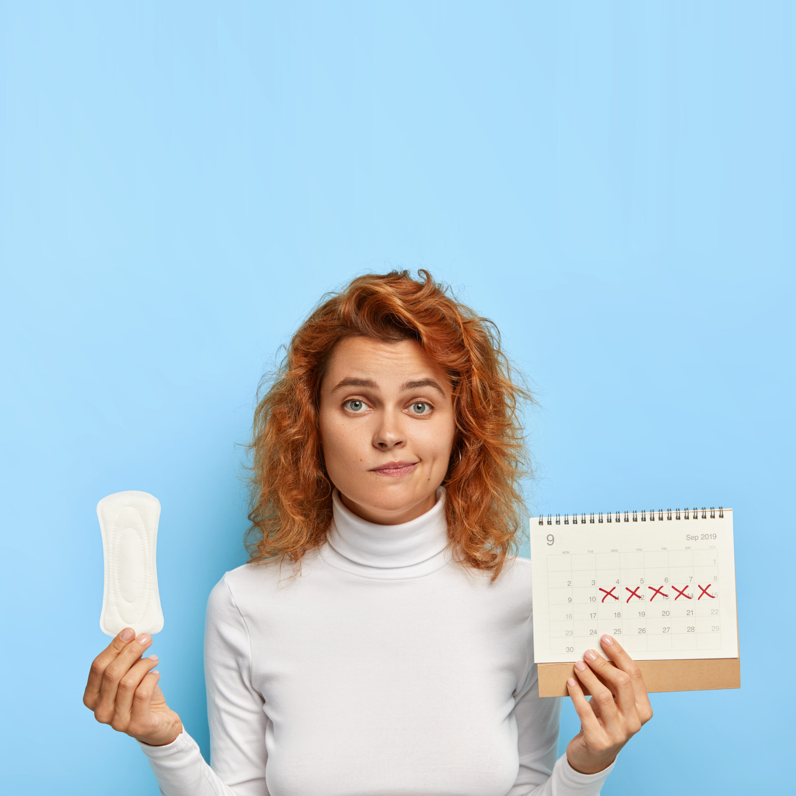 Puzzled ginger woman holds sanitary napkin and menstruation calendar with marked red days, suffers from period cramps, controls her women health, has periods this week, poses over blue background